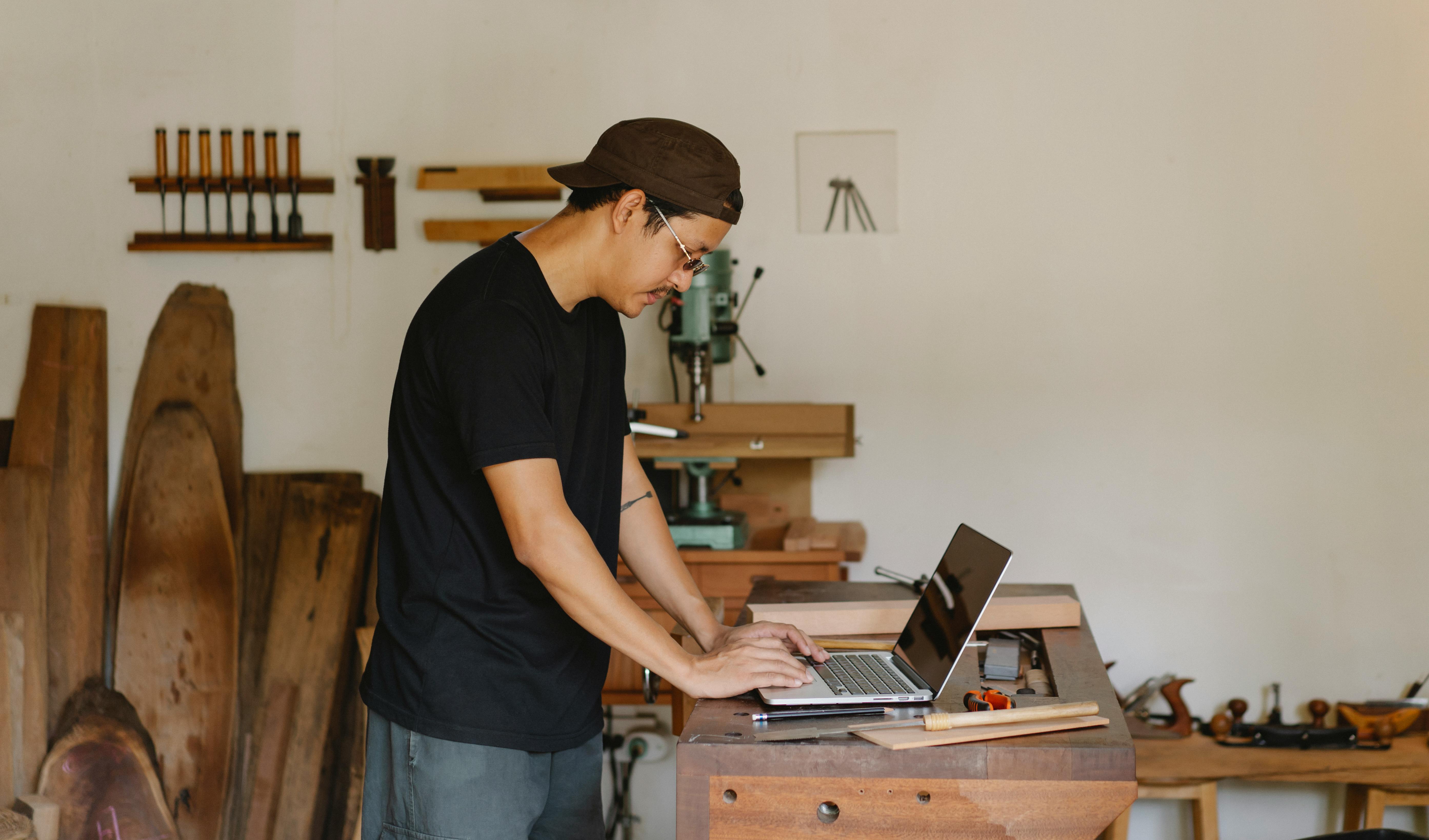 Side view of young ethnic male designer in casual clothes and eyeglasses typing on laptop while working in carpentry workshop